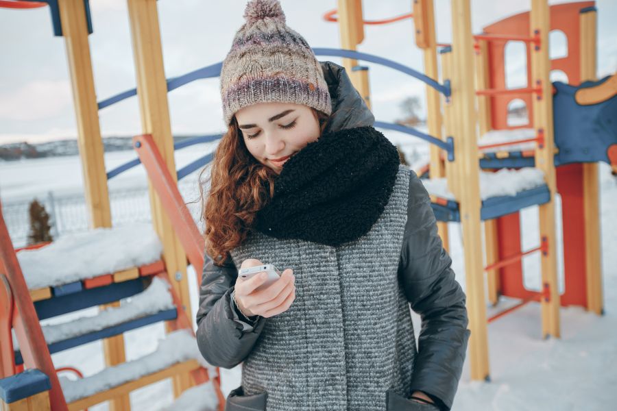 Young woman in winter clothes checking her phone in a snowy playground, symbolizing use of a snow day calculator app.